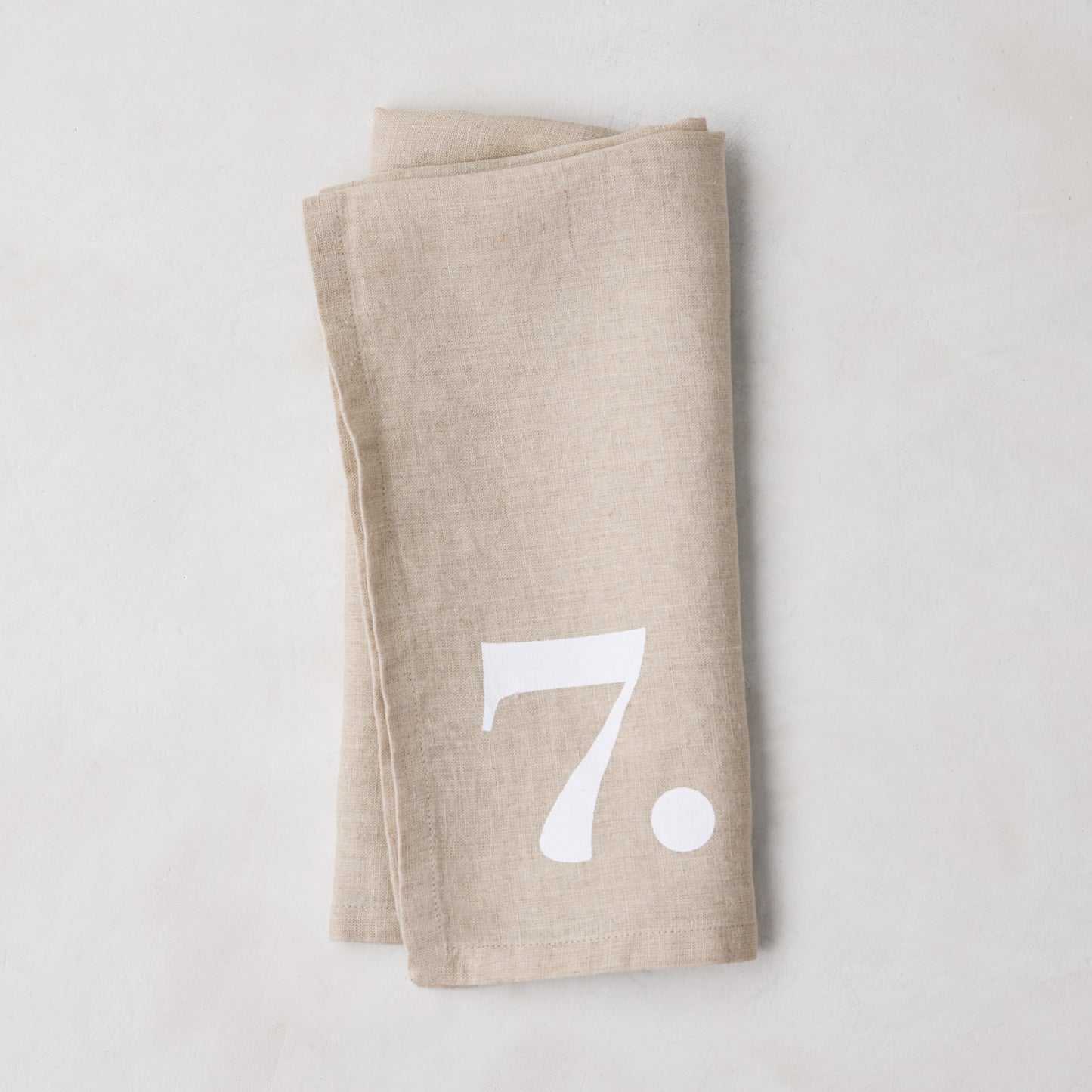 Diane Keaton + Hudson Grace Khaki Linen Numeric Napkin, folded, with a white number 7.