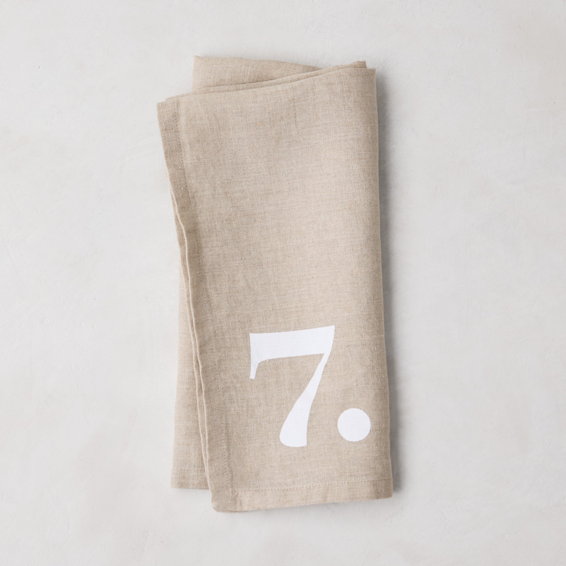 Diane Keaton + Hudson Grace Khaki Linen Numeric Napkin, folded, with a white number 7.