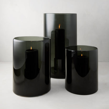 Three Diane Keaton x Hudson Grace smoke grey glass hurricanes with lit black pillar candles.