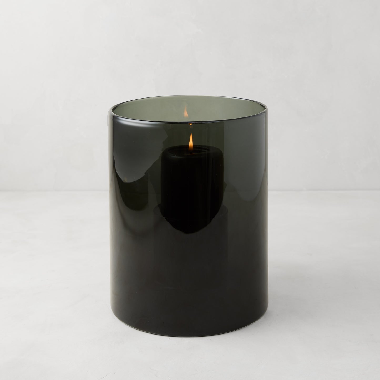 Small Smoke Grey Glass Hurricane from Diane Keaton + Hudson Grace, with a lit black pillar candle inside.