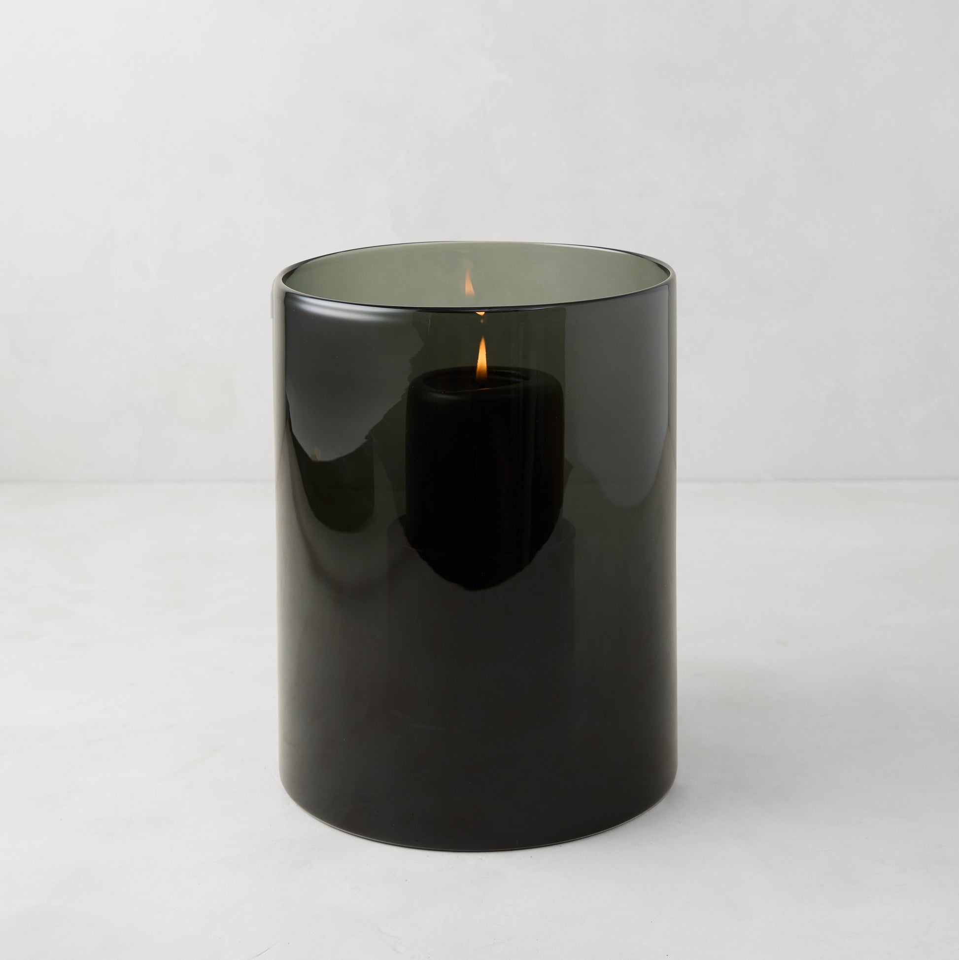 Small Smoke Grey Glass Hurricane from Diane Keaton + Hudson Grace, with a lit black pillar candle inside.