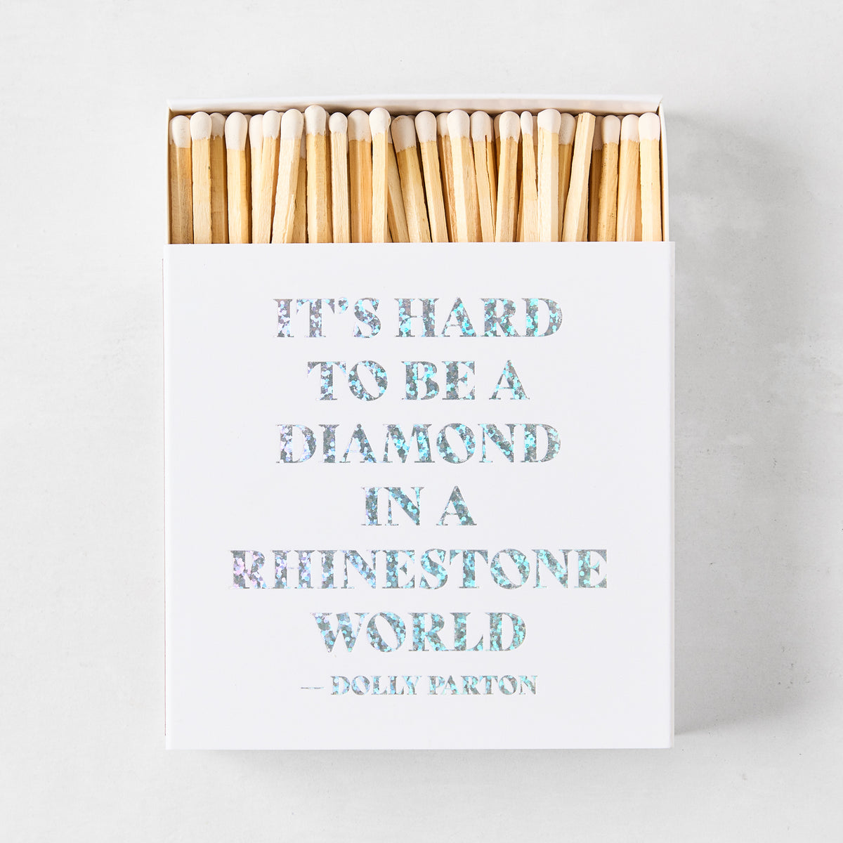 Square white matchbox featuring holographic text reading It's Hard to Be a Diamond in a Rhinestone World by Dolly Parton.