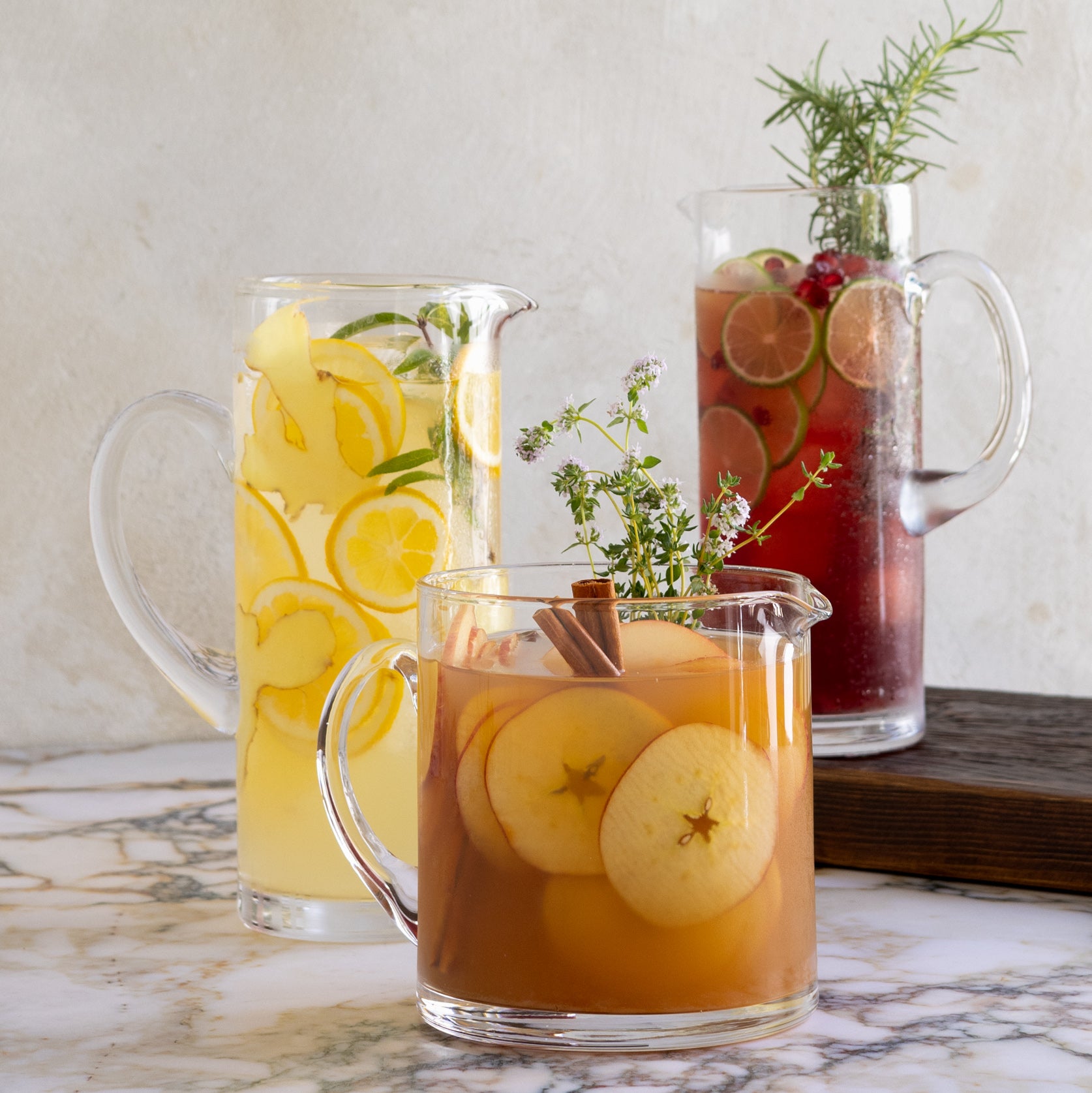 Three clear glass Downtown Tall Bar Pitchers, filled with colorful drinks and garnishes, on a marble surface.