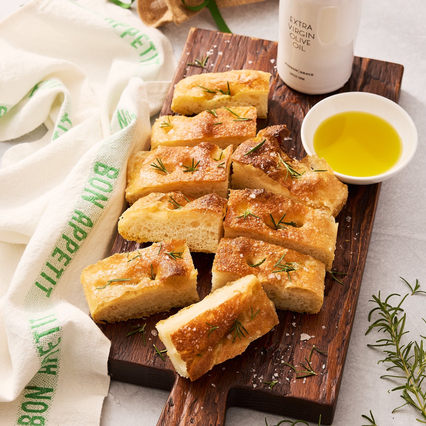Golden focaccia bread slices with rosemary and sea salt on a dark wood board, with olive oil.