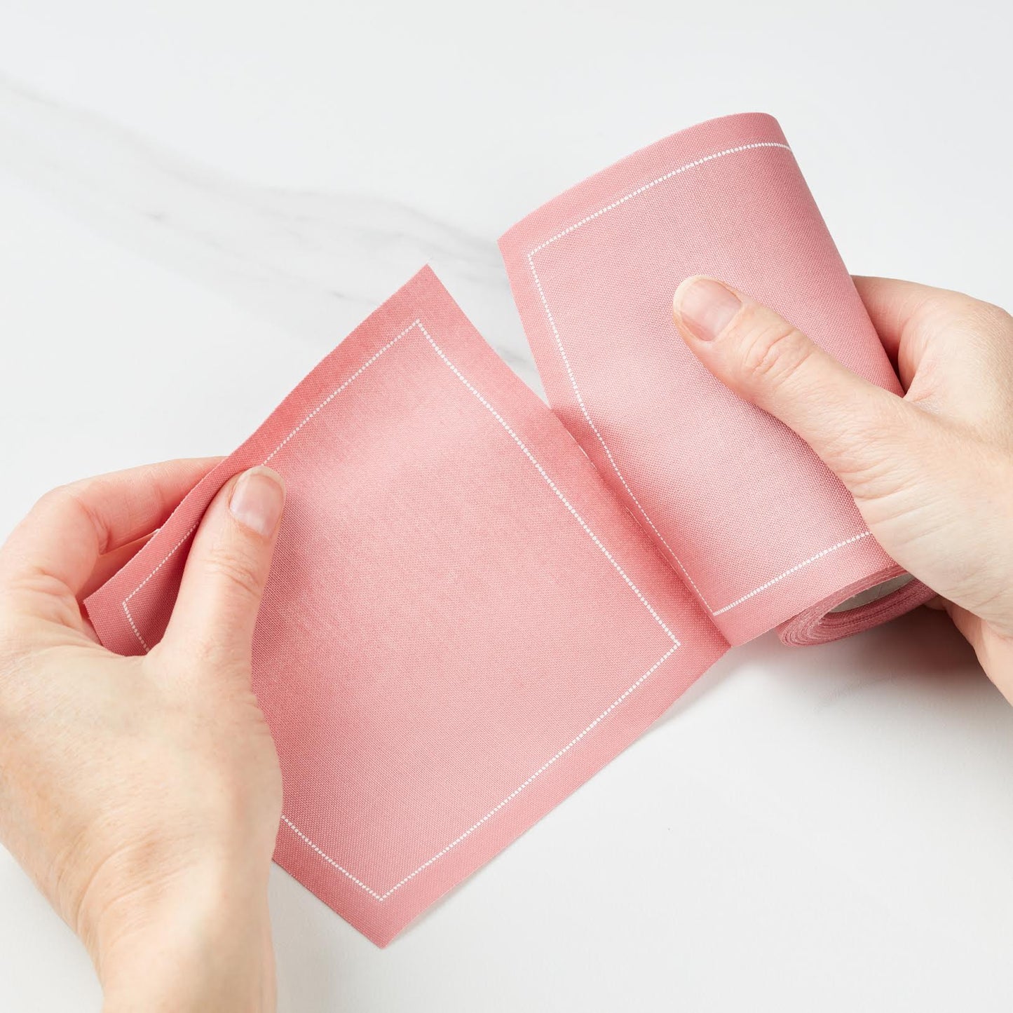 Hands tearing a dusty pink cotton cocktail napkin from a roll, showing white stitched borders.