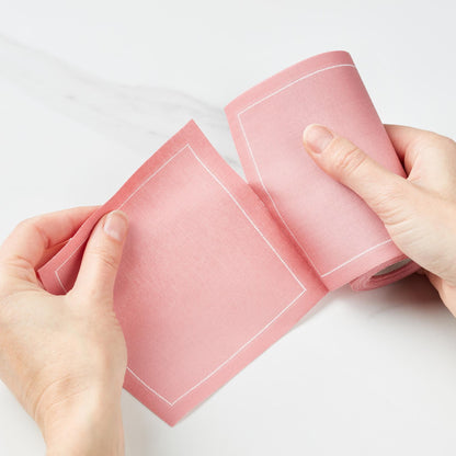 Hands tearing a dusty pink cotton cocktail napkin from a roll, showing white stitched borders.
