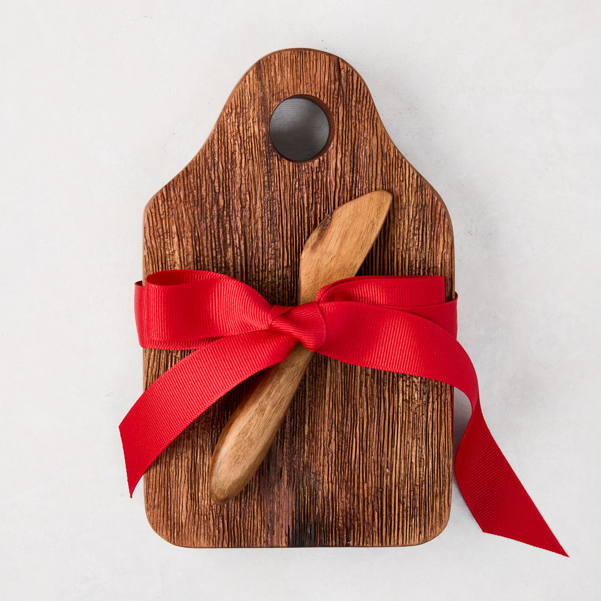 Acacia wood cheese board with a small hole, natural wood grain, and a wooden spreader, tied with a red bow.