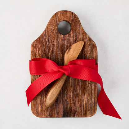 Acacia wood cheese board with a small hole, natural wood grain, and a wooden spreader, tied with a red bow.