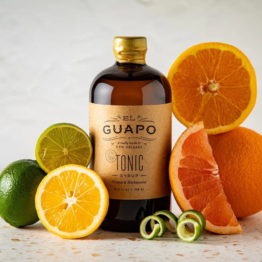 El Guapo Tonic Syrup in an amber bottle with a kraft label, surrounded by fresh limes, lemons, oranges, and grapefruit.