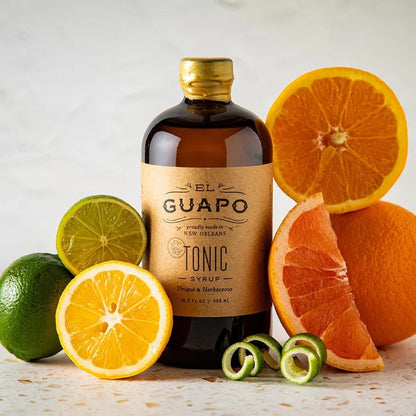 El Guapo Tonic Syrup in an amber bottle with a kraft label, surrounded by fresh limes, lemons, oranges, and grapefruit.