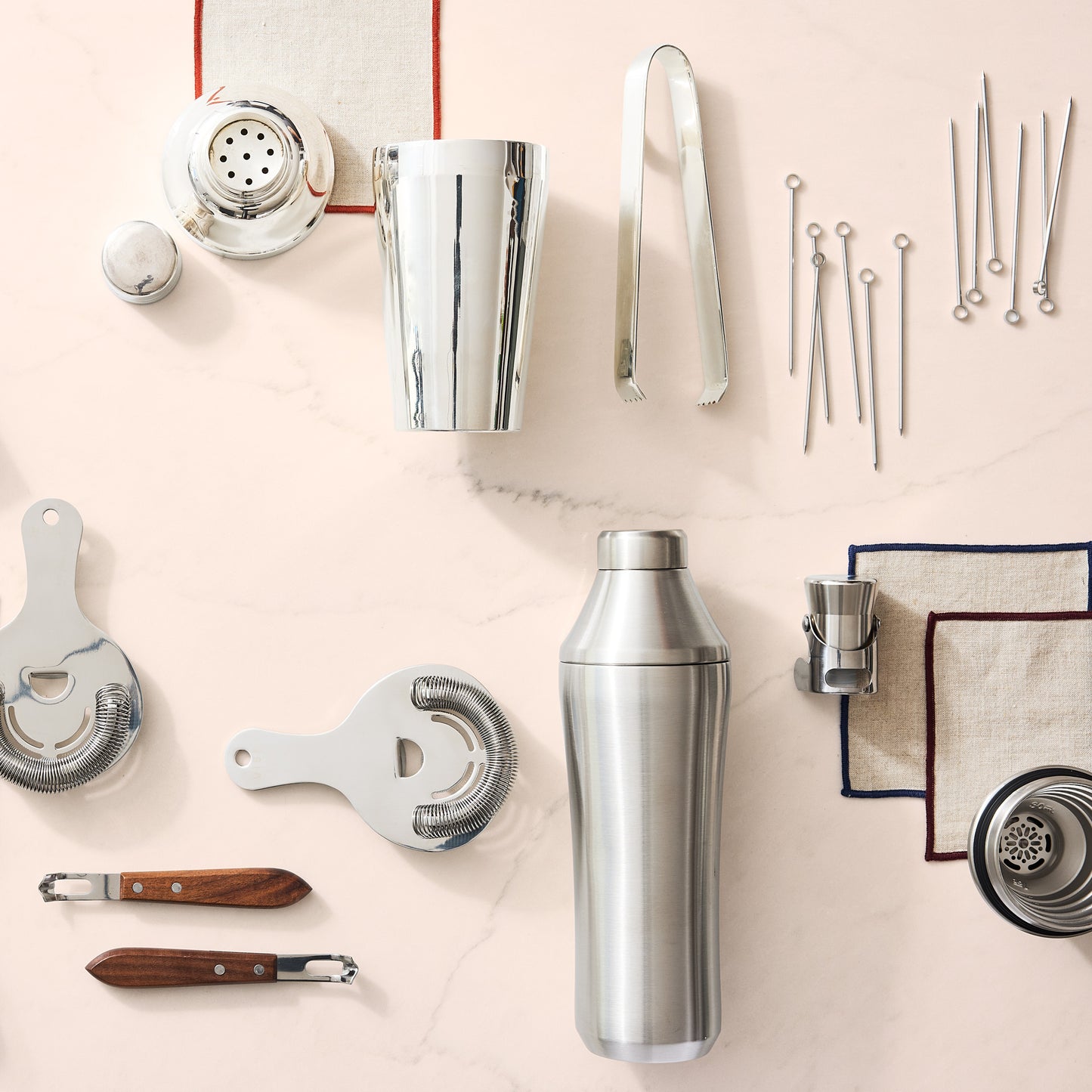 Silver stainless steel craft cocktail shaker set with strainers, jigger, tongs, and picks on a light marble surface.