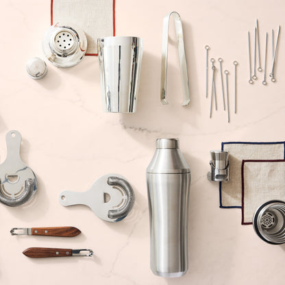 Silver stainless steel craft cocktail shaker set with strainers, jigger, tongs, and picks on a light marble surface.