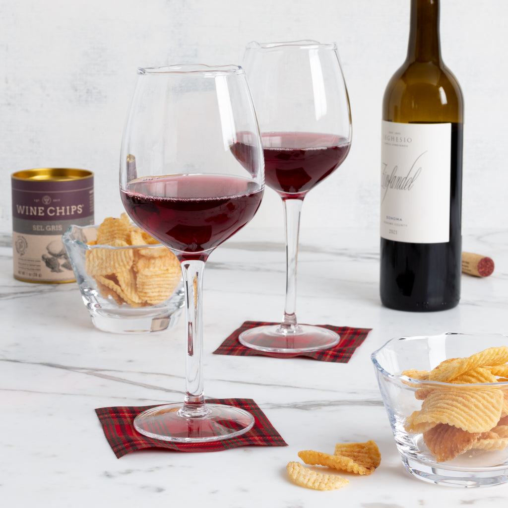 Two Elijah Large Scissor-Cut Wine Glasses filled with red wine, on red plaid coasters, with chips and a wine bottle.