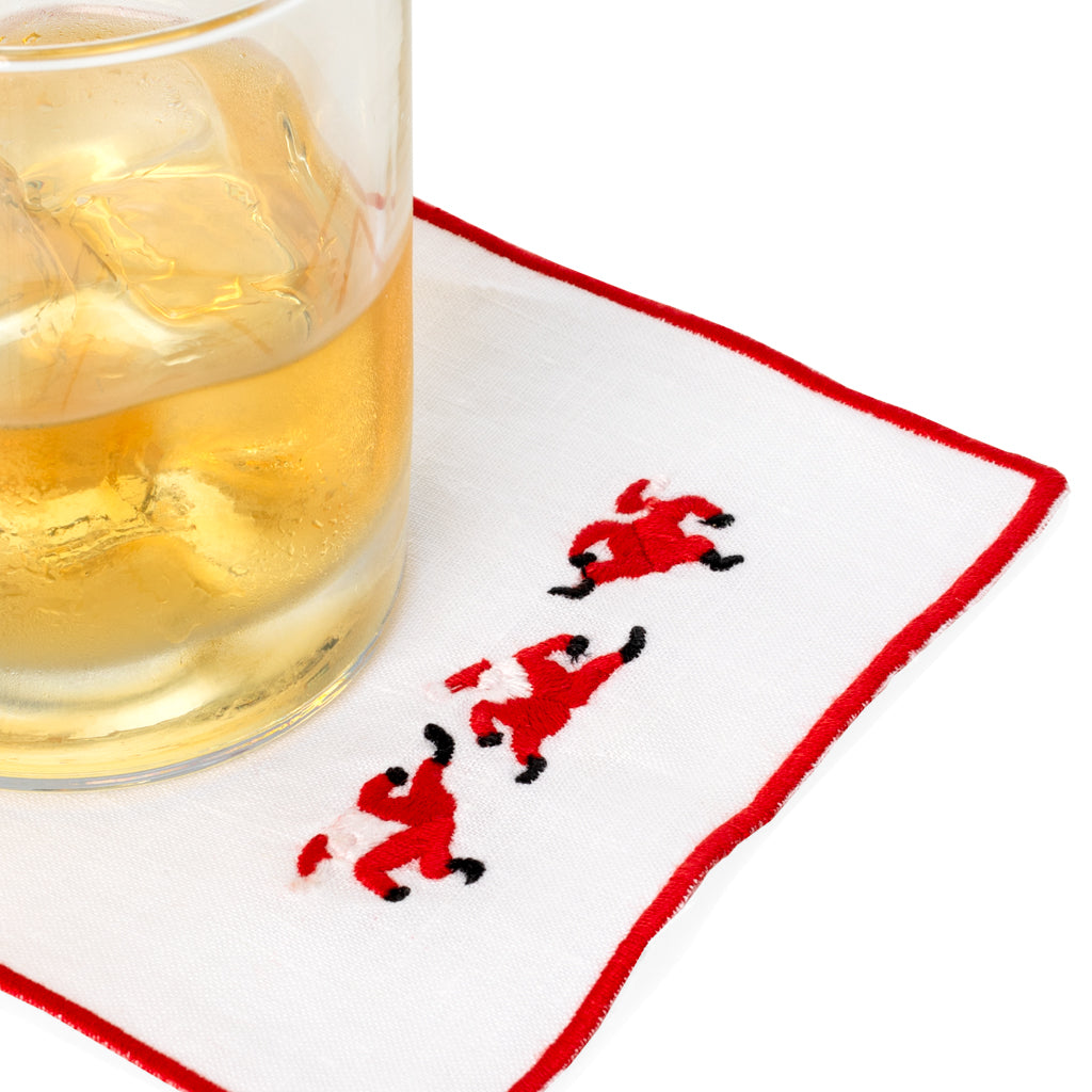 White linen coaster with red embroidered Santa figures and a red border, under a glass with ice.