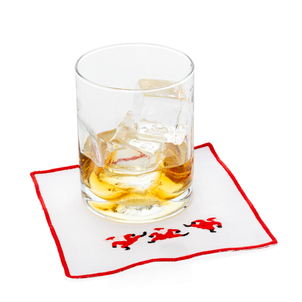 White linen coaster with red embroidered Santa figures and red border, under a glass of whiskey.