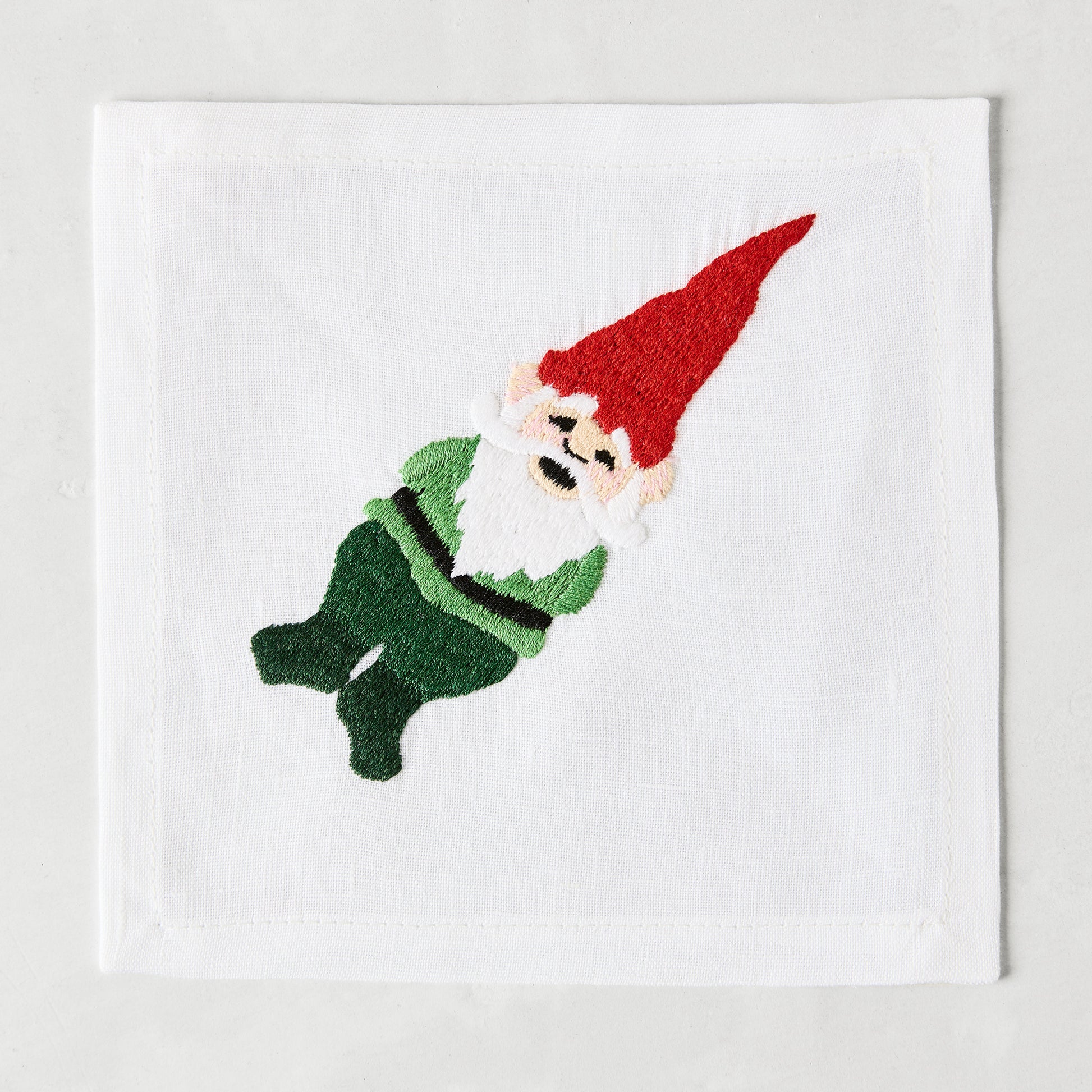 White linen cocktail napkin coaster with embroidered gnome in red hat and green outfit.