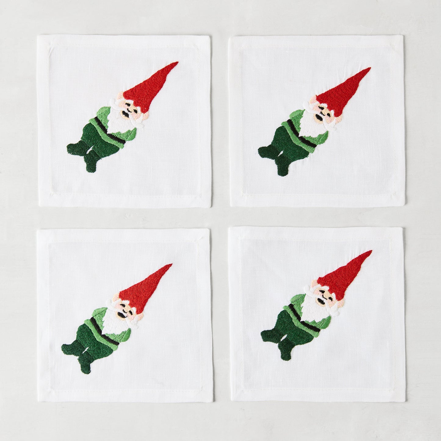 Set of 4 white linen 5.5" square cocktail napkins with embroidered gnomes in red hats and green outfits.
