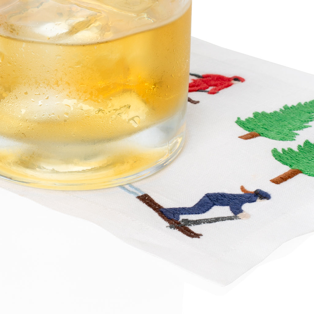 White linen coaster with embroidered skiers and green trees, under a glass of iced drink.