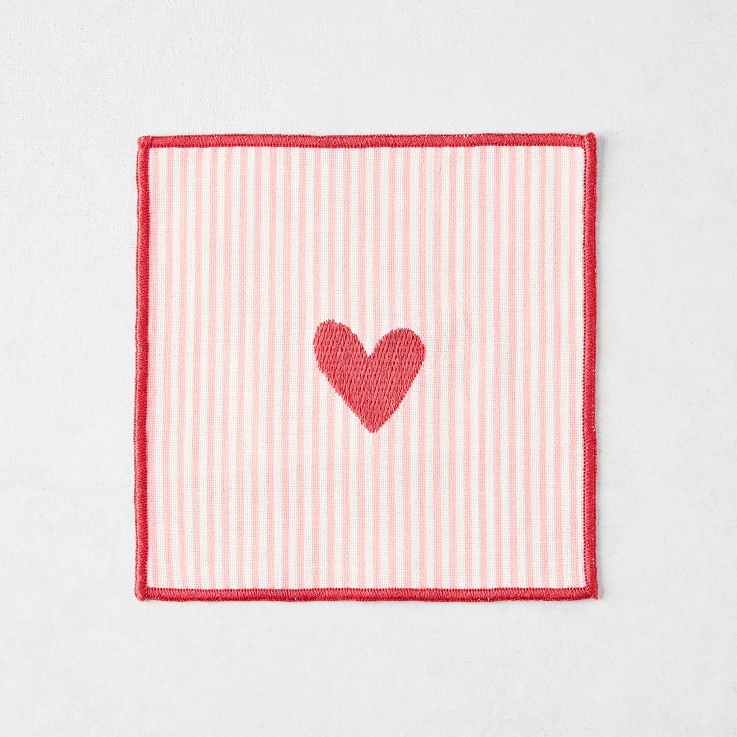 Cotton coaster with pale pink and white stripes, red embroidered heart, and red edge.