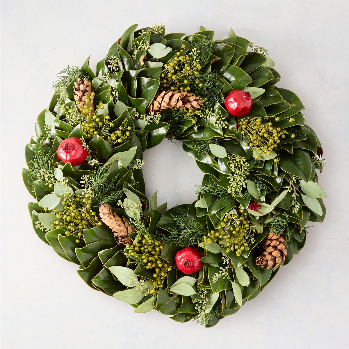 24-inch holiday wreath featuring magnolia leaves, fir, boxwood, pinecones, and red pomegranates on a light background.