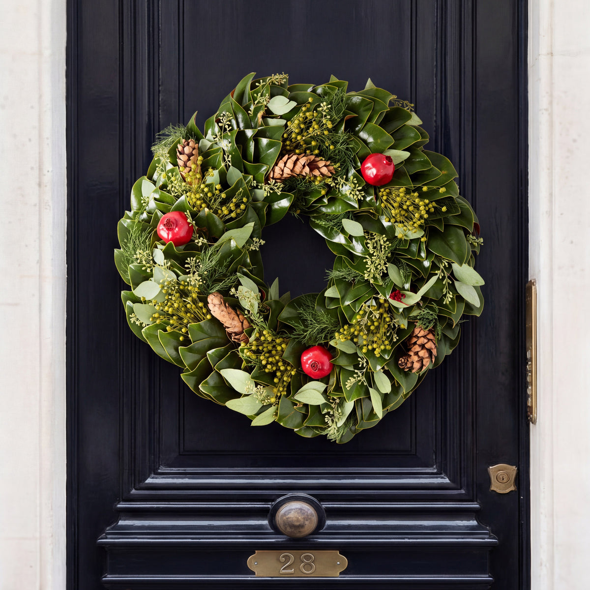 24-inch holiday wreath featuring magnolia leaves, boxwood, fir, pinecones, and red pomegranates on a black door.