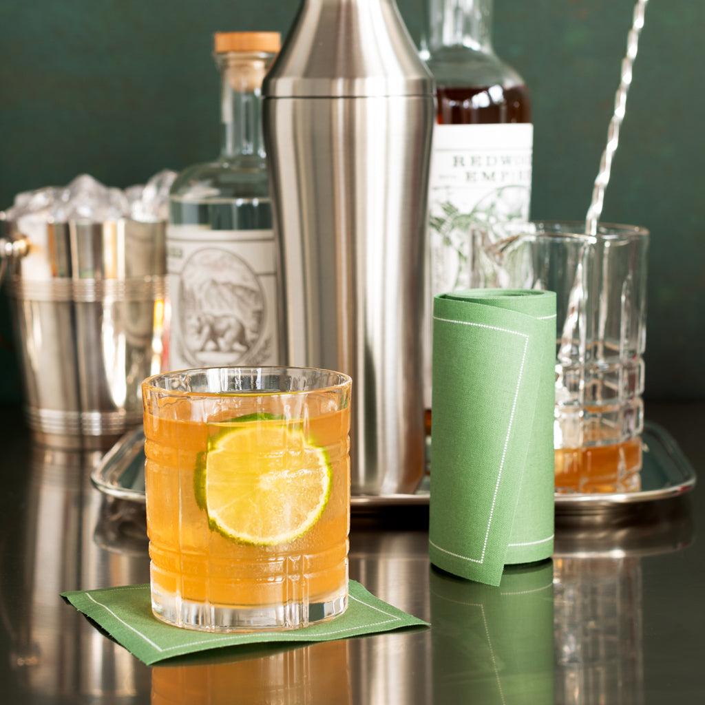 Green eucalyptus tear-off cotton cocktail napkin under a drink, with a rolled napkin nearby.