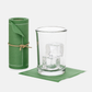 Roll of 50 eucalyptus green cotton cocktail napkins with white stitching, next to a glass with ice on a single napkin.