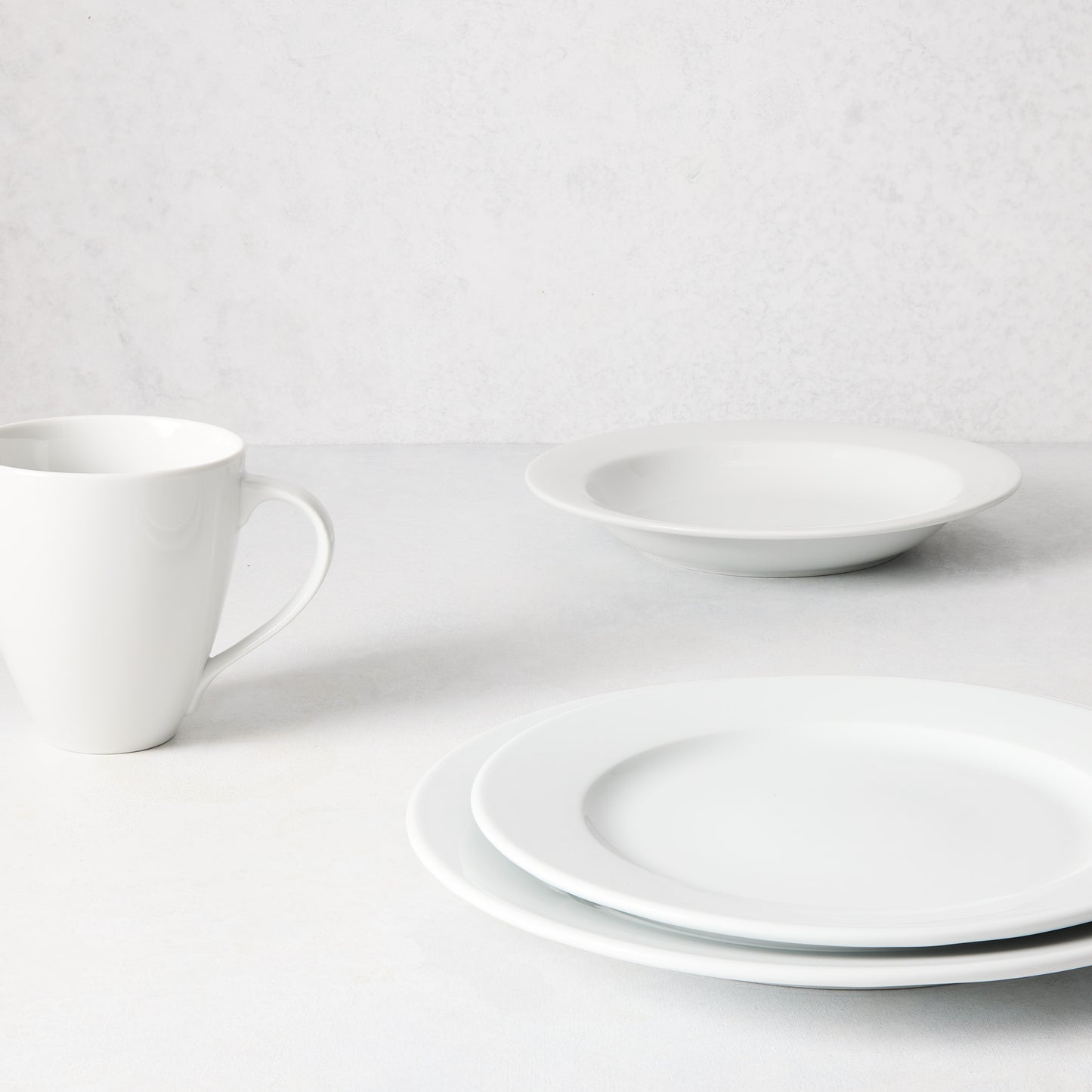 White porcelain Everyday Bowl, dinner plate, salad plate, and mug on a light background.