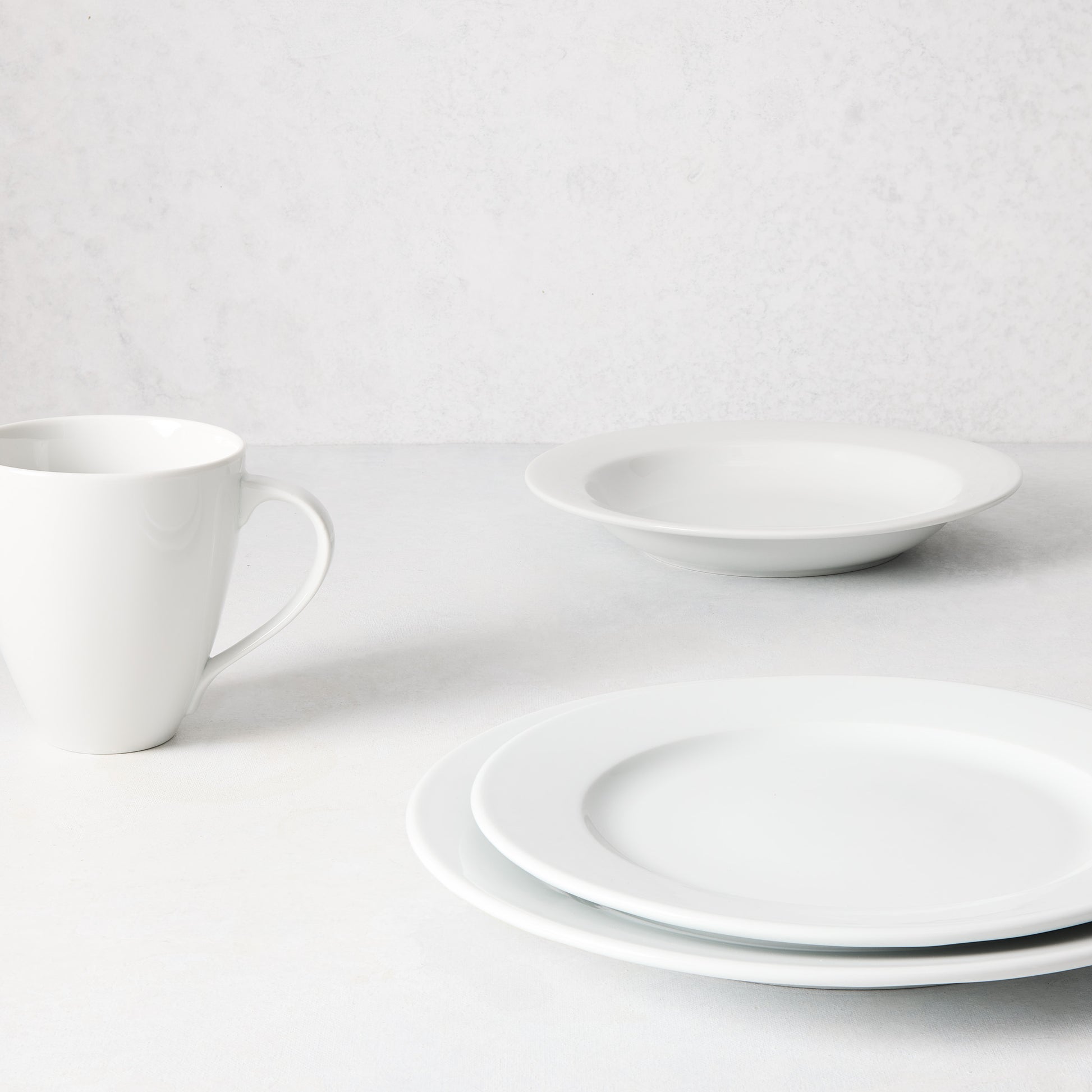 White porcelain Everyday Bowl, dinner plate, salad plate, and mug on a light background.