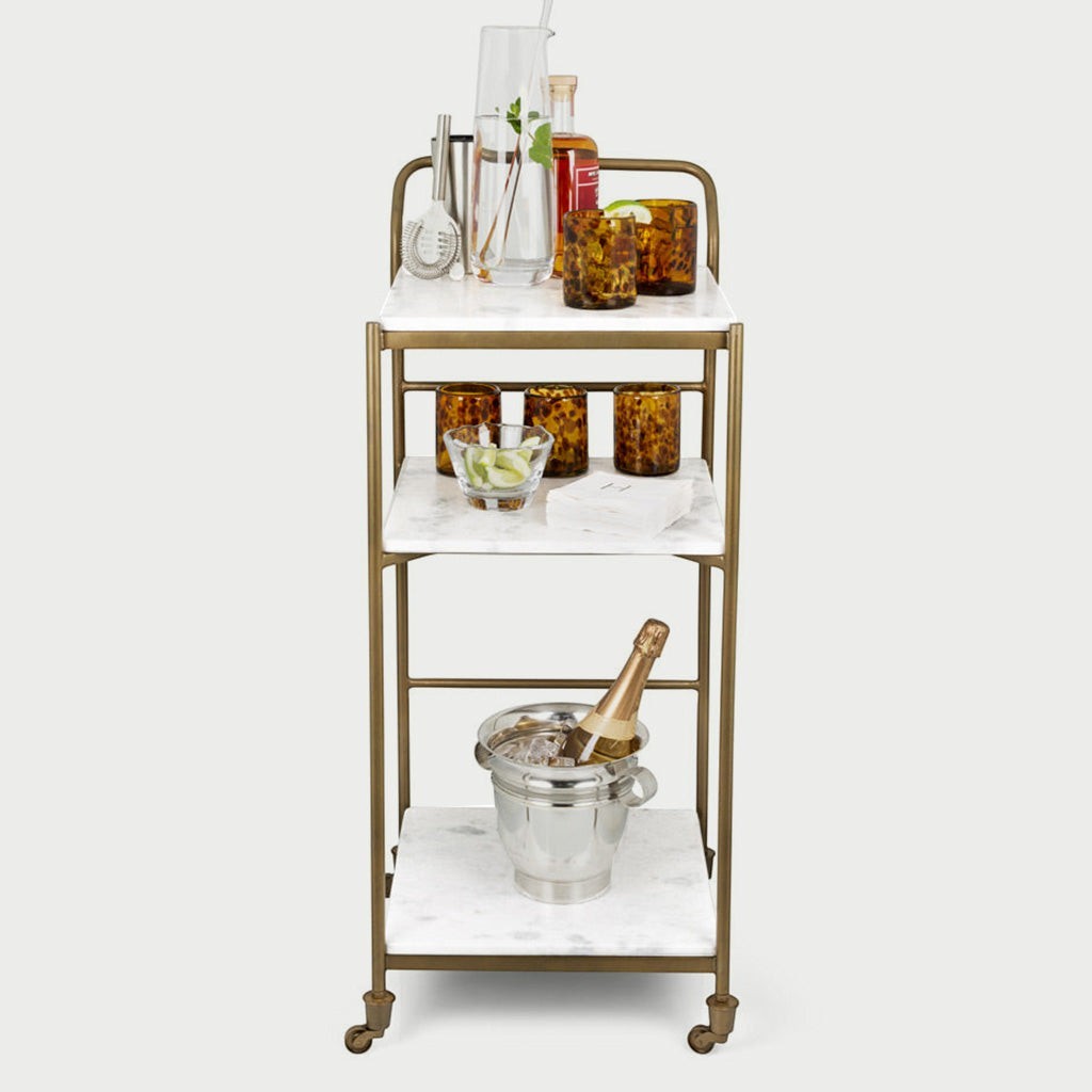 Felix antique brass bar cart with three white marble shelves, styled with barware and drinks.