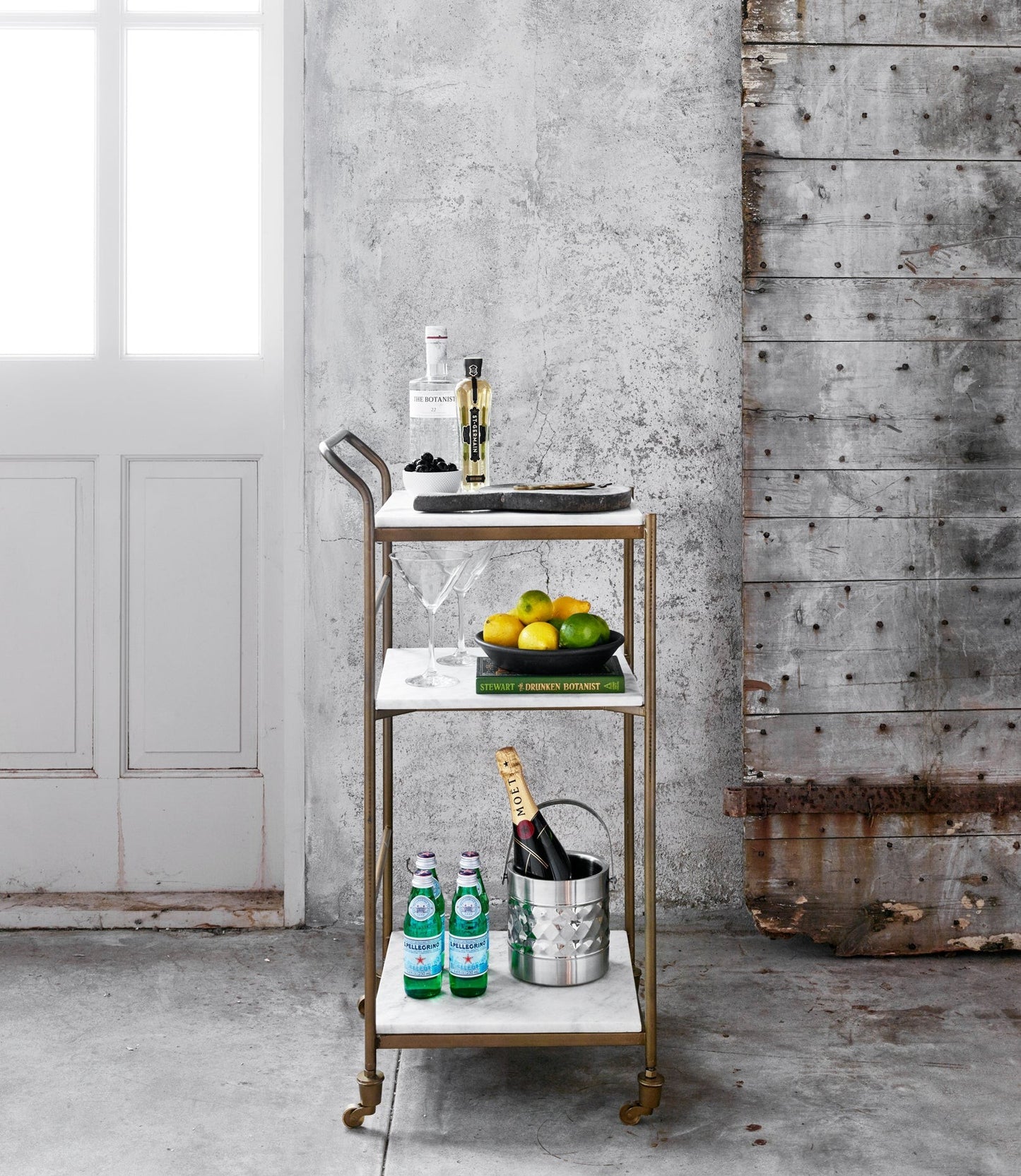 Felix antique brass and white marble square bar cart with three shelves, bottles, and fruit.