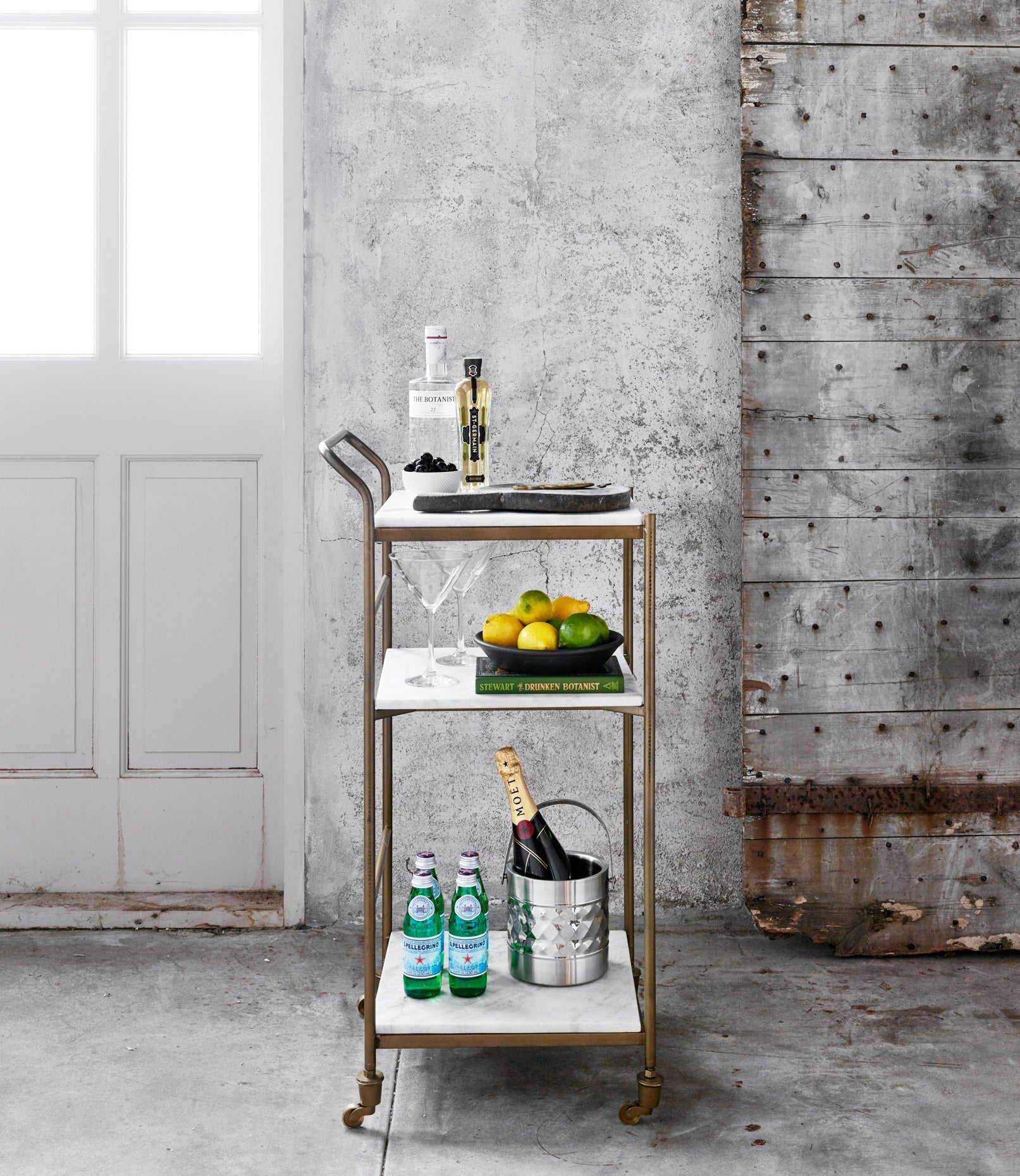 Felix antique brass and white marble square bar cart with three shelves, bottles, and fruit.