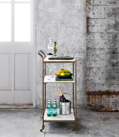 Felix antique brass and white marble square bar cart with three shelves, bottles, and fruit.