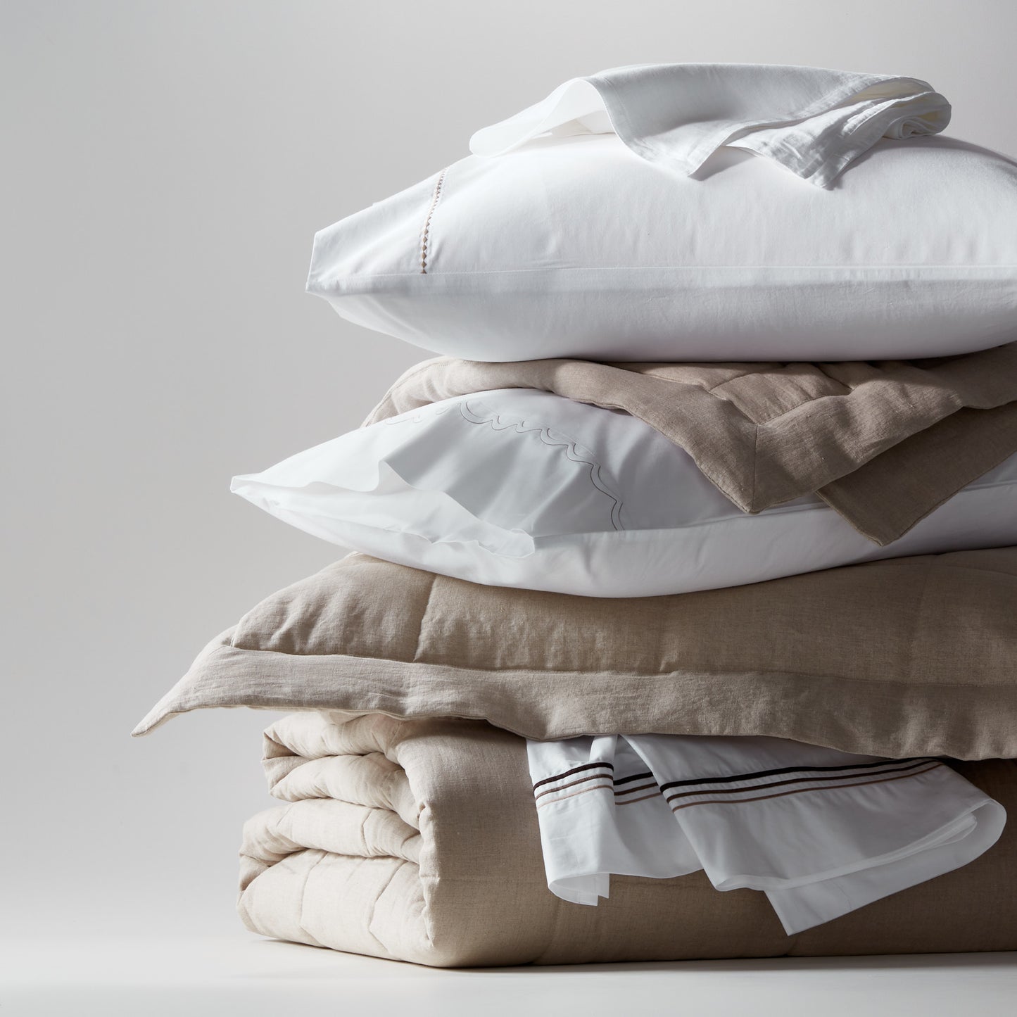 Stack of flax linen and cotton bedding: white pillows, natural linen quilted coverlet, and sheets.