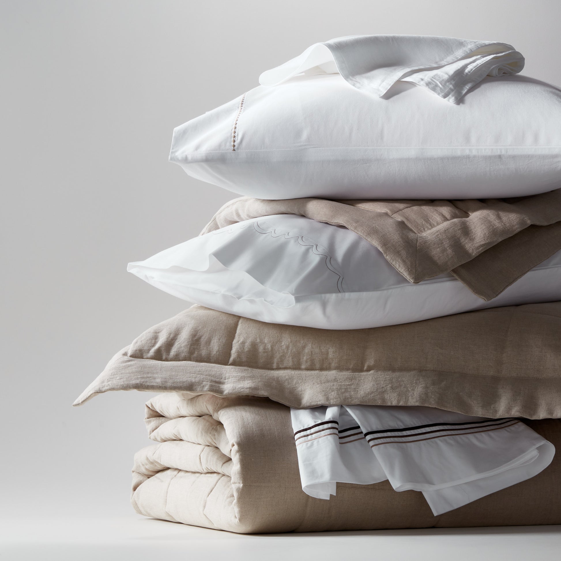 Stack of flax linen and cotton bedding: white pillows, natural linen quilted coverlet, and sheets.