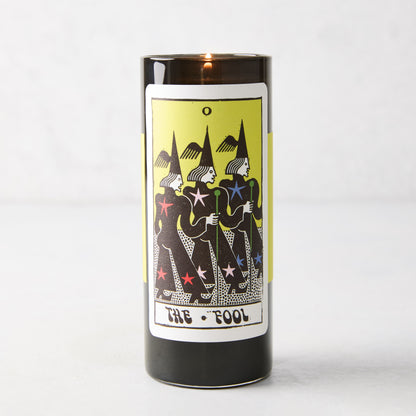 Tall black glass pillar candle featuring a yellow The Fool tarot card illustration with three figures and stars.