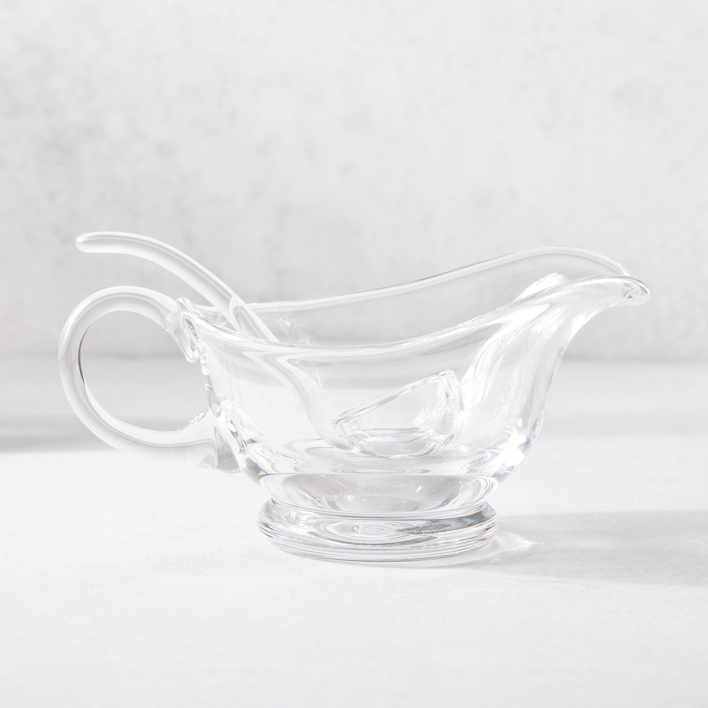 Clear glass gravy boat with handle, spout, and matching ladle on a white surface.