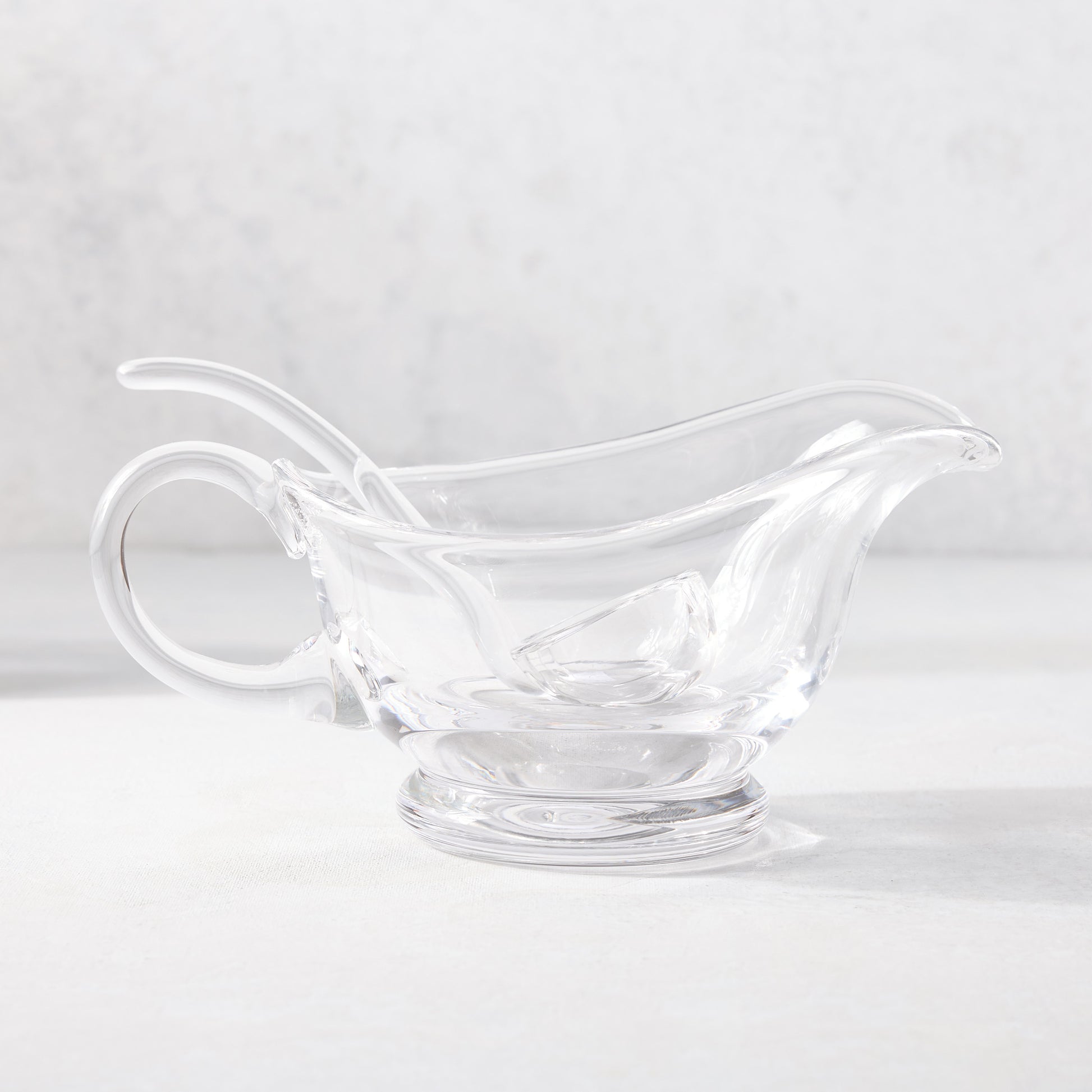 Clear glass gravy boat with handle, spout, and matching ladle on a white surface.