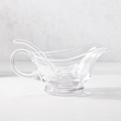 Clear glass gravy boat with handle, spout, and matching ladle on a white surface.