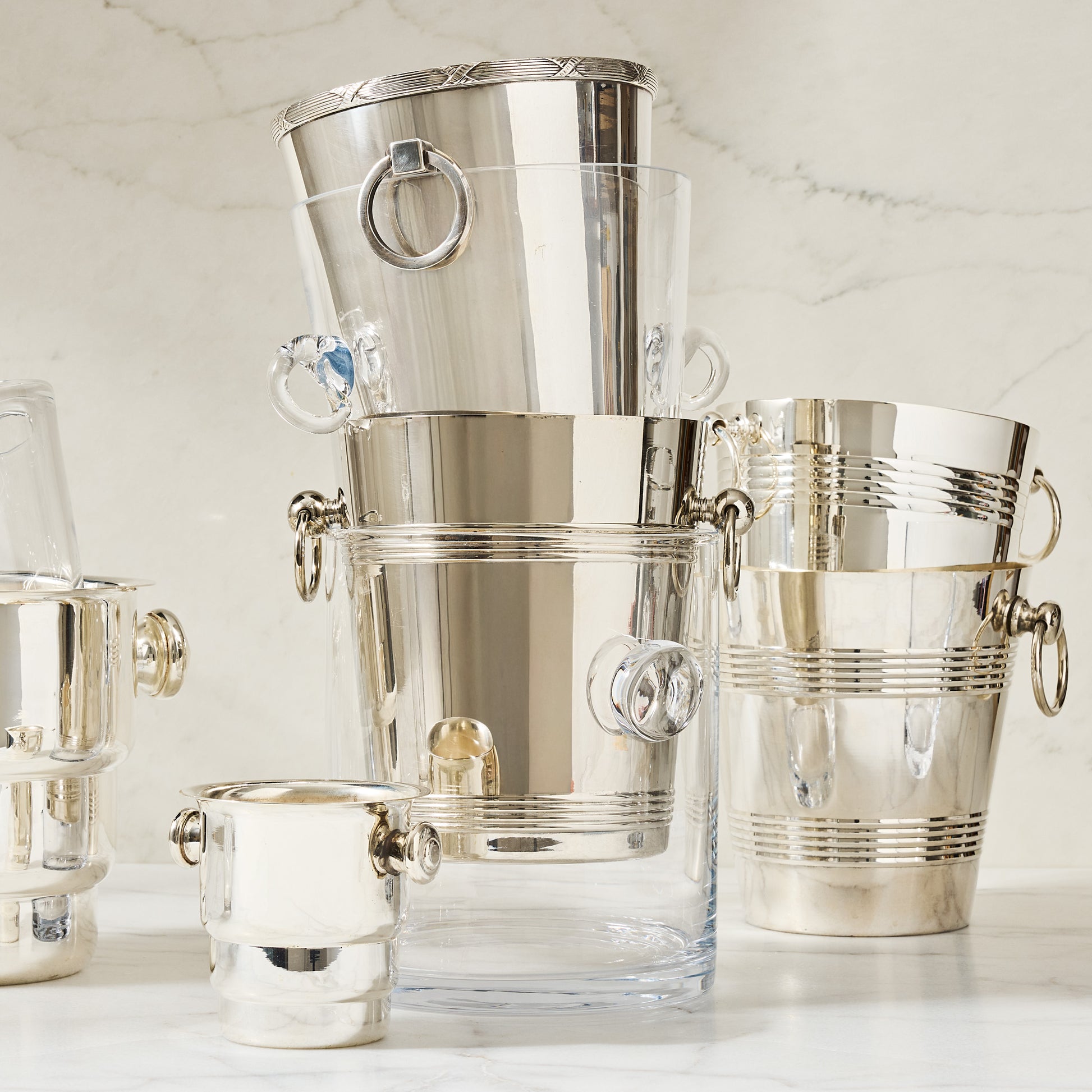 Stacked glass and silver ice buckets with ring handles on a marble surface.
