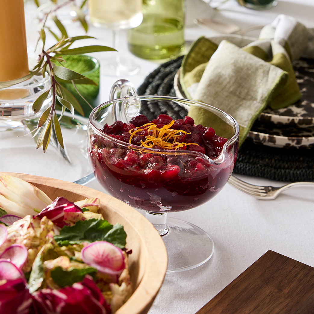 Clear glass gravy boat with handle, filled with cranberry sauce and orange zest, on a white tablecloth.