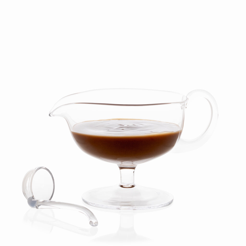 Clear glass sauce/gravy boat with handle, filled with brown gravy, and a small glass ladle.