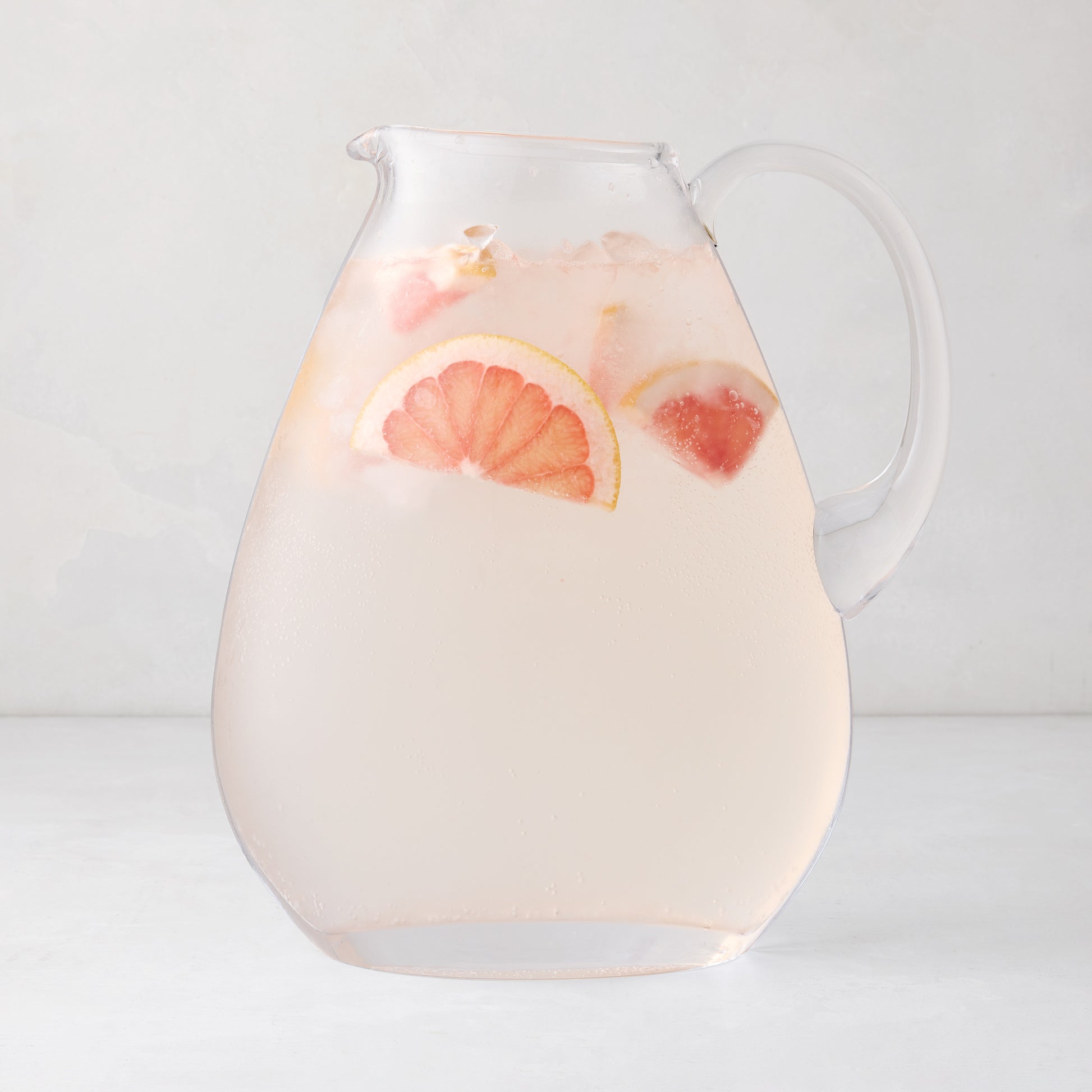 Clear glass teardrop pitcher filled with pink grapefruit drink and ice.