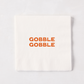 Square white paper cocktail napkin with orange Gobble Gobble text, 5.5 inches.