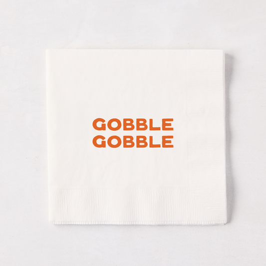 Square white paper cocktail napkin with orange Gobble Gobble text, 5.5 inches.