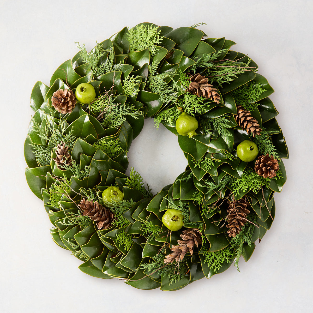 24-inch wreath featuring fresh green magnolia leaves, cedar branches, pinecones, and pomegranates on a white background.