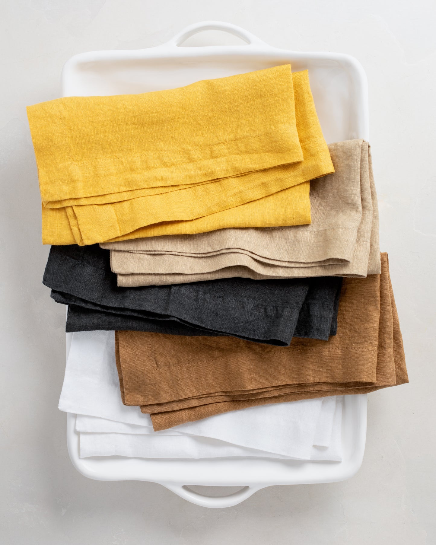 Stack of five folded 22" square linen napkins in golden mustard, beige, charcoal, brown, and white.
