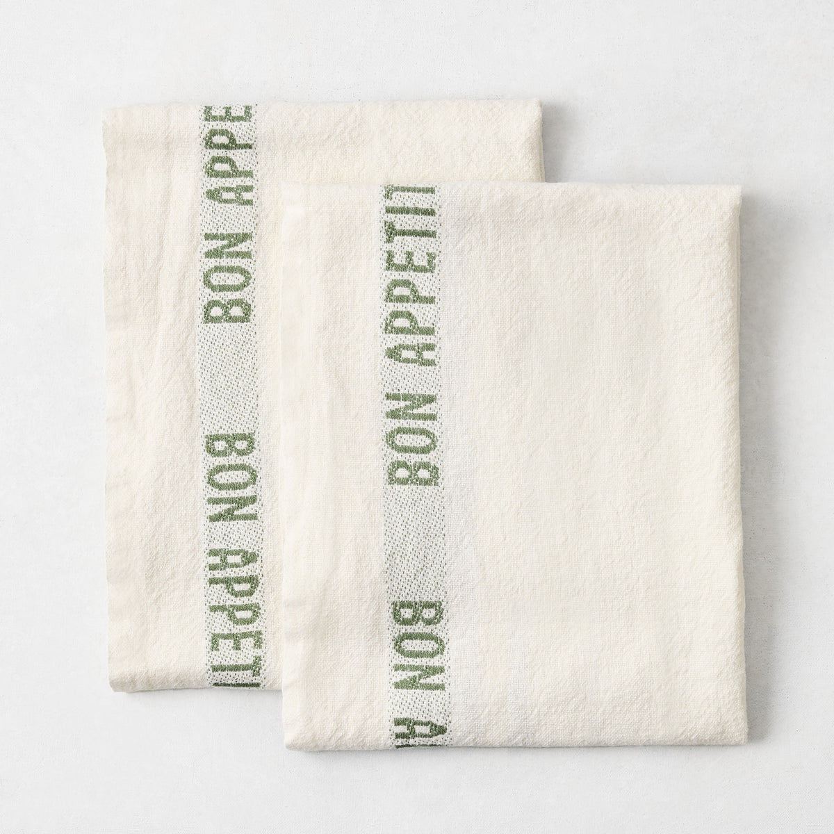 Set of two folded, off-white 100% linen French tea towels with green "Bon Appetit" text and striped detailing.