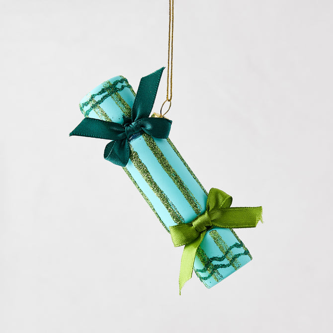 Light blue Christmas cracker ornament with green glitter stripes and dark green ribbon bows hanging on a gold string.