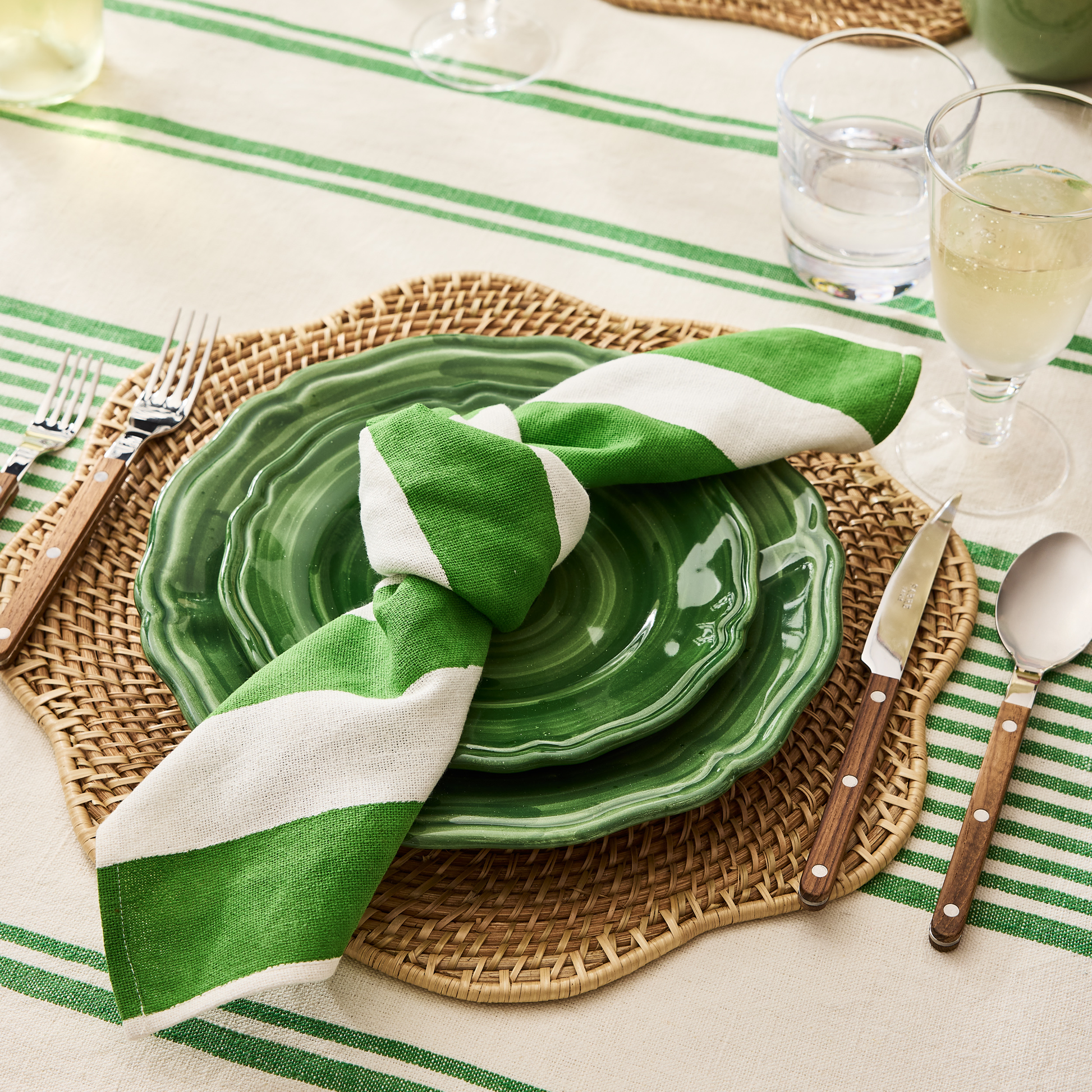 Green Hampton Cotton Stripe Napkin, tied on a green ceramic plate, on a woven placemat.